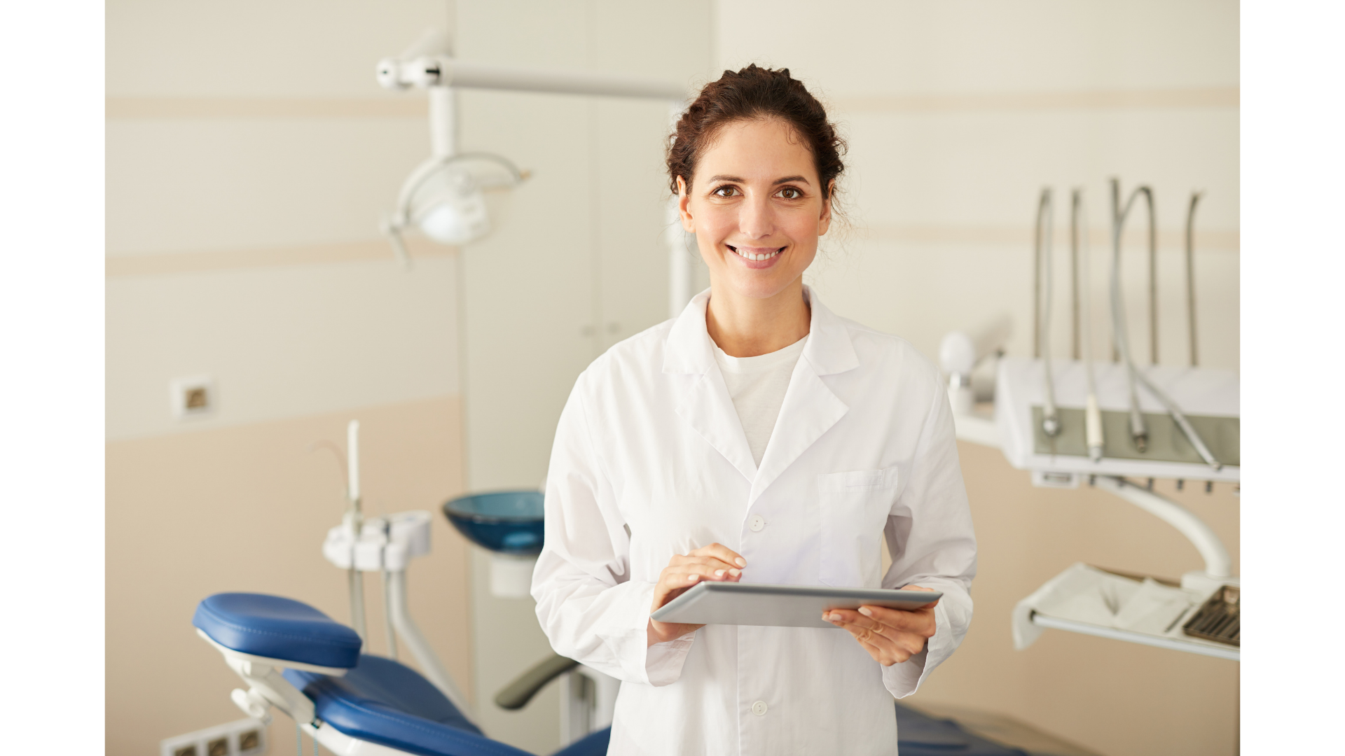 Dentista sorridente de jaleco branco segurando um tablet, em consultório odontológico claro